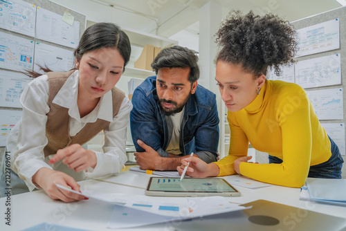 Three colleagues lean over a desk covered in printed charts and a tablet, closely reviewing data together during a focused analysis session in a modern office lined with project boards.