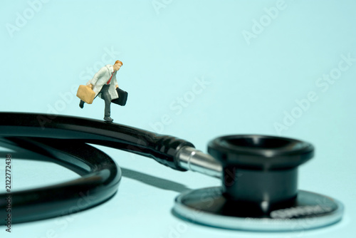 Miniature tiny people toy figure photography. A men doctor running above black stethoscope. Isolated on blue background.