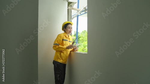 Home inspector in yellow hard hat and safety jacket checking house interior near window opening, inspecting wall repair and room renovation with tablet in bright home workspace