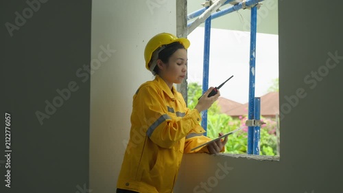Home inspector woman in yellow hard hat and jacket using walkie talkie near window at unfinished building site, checking interior through opening with clipboard and safety gear