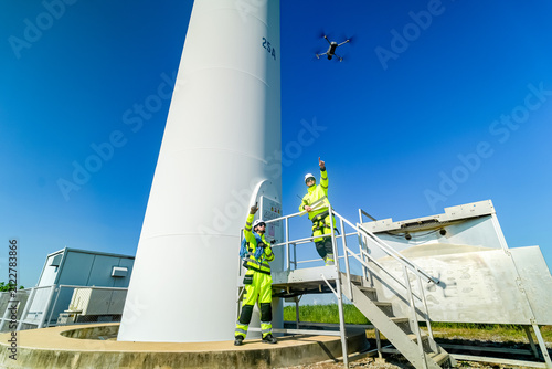 Workers in safety gear operate a drone near a tall wind turbine in a clear sky during daytime