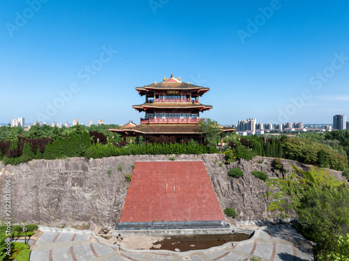 Traditional Chinese pavilion with golden roof stands atop rocky cliff with red staircase and plaza under clear blue sky