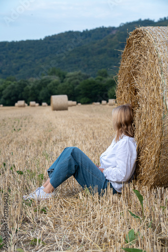 Unrecognizable Woman Sitting Against Round Hay Bale in Harvested Field with Rural Landscape