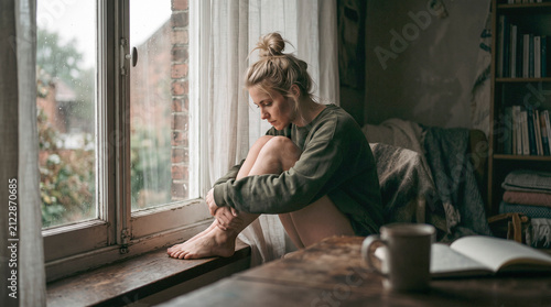 Young woman sitting on a windowsill in a cozy home, hugging her knees and looking down in quiet reflection. Soft natural light, calm interior, solitude, introspection, emotional well-being, lifestyle.
