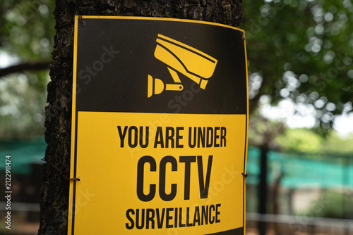Konstfotografi Yellow CCTV Surveillance Warning Sign Tied to Tree in Outdoor Public Park