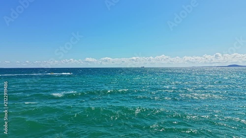 Waves gently roll on the surface of the ocean under a bright blue sky. Clouds linger in the distance while a ship can be seen far off at sea.