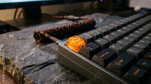 Keyboard mechanical components feature a customized orange escape key and a braided cable resting upon a dark slate surface, emphasizing the tactile precision of high-end custom computer peripherals.