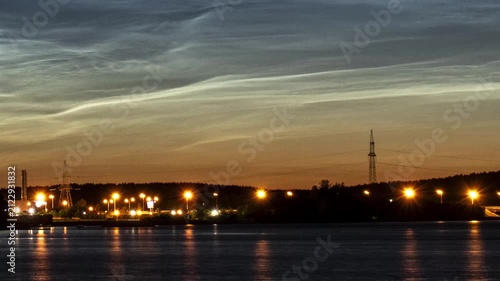Noctilucent clouds glowing over a river with city lights and a bridge at twilight
