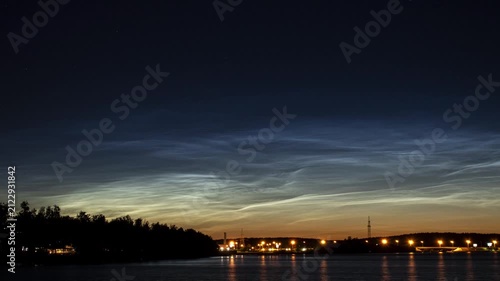 Noctilucent clouds glowing over a river with city lights and a bridge at twilight
