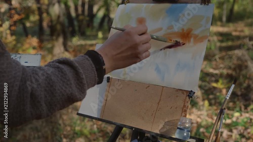The artist paints an autumn landscape in watercolor while standing at an easel in the park