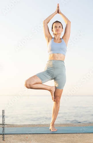 Konstfotografi Sedulous woman doing warrior one leg pose of yoga on mat on beach at sunrise out