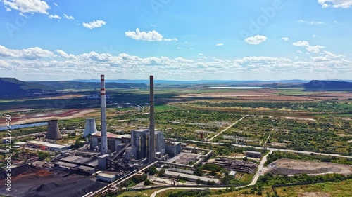 A large power plant stands in an industrial zone next to a river. Fields and hills surround the site. The sky has some clouds and looks clear. It is daytime.