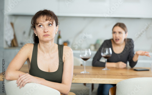 Konstfotografi Sad middle-aged woman sitting at table and other woman brawling to her behind in