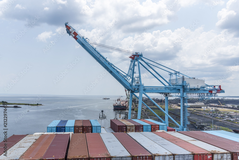Naklejka premium Large container vessel alongside a busy container port with loading crane and stacked shipping containers, with visible entrance to the port of Mobile, Alabama, USA.