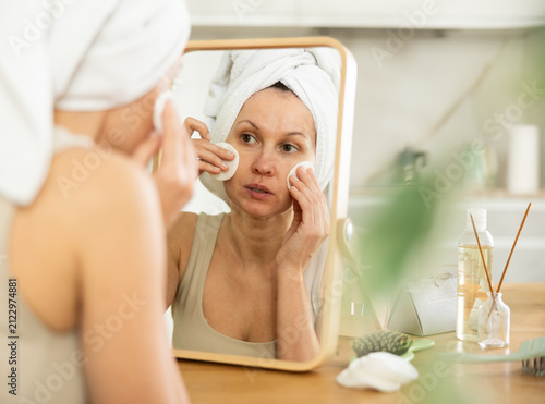 фотографія Attentive woman in her middle age using cotton pads to purify her face looking i