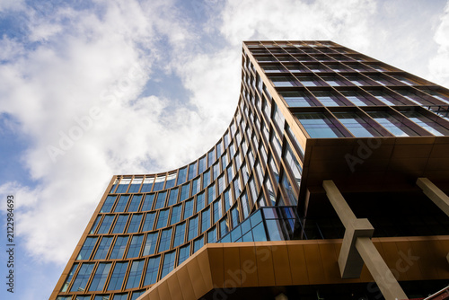 Dynamic low angle view of a curved contemporary office tower in Brussels featuring bronze framed glass architecture, dramatic clouds and innovative European business design with generous copy space.