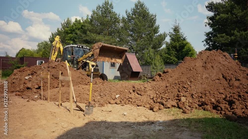 Heavy construction machinery working on a building site, with an excavator digging soil and moving earth to prepare the ground for foundation work during a sunny day with blue sky and trees