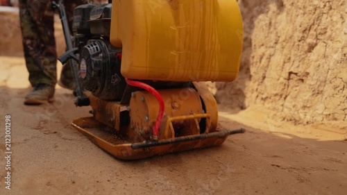 Construction worker operating a vibratory plate compactor to level and compact sand, creating a solid foundation for paving during roadworks, with dust and grains flying from the vibration
