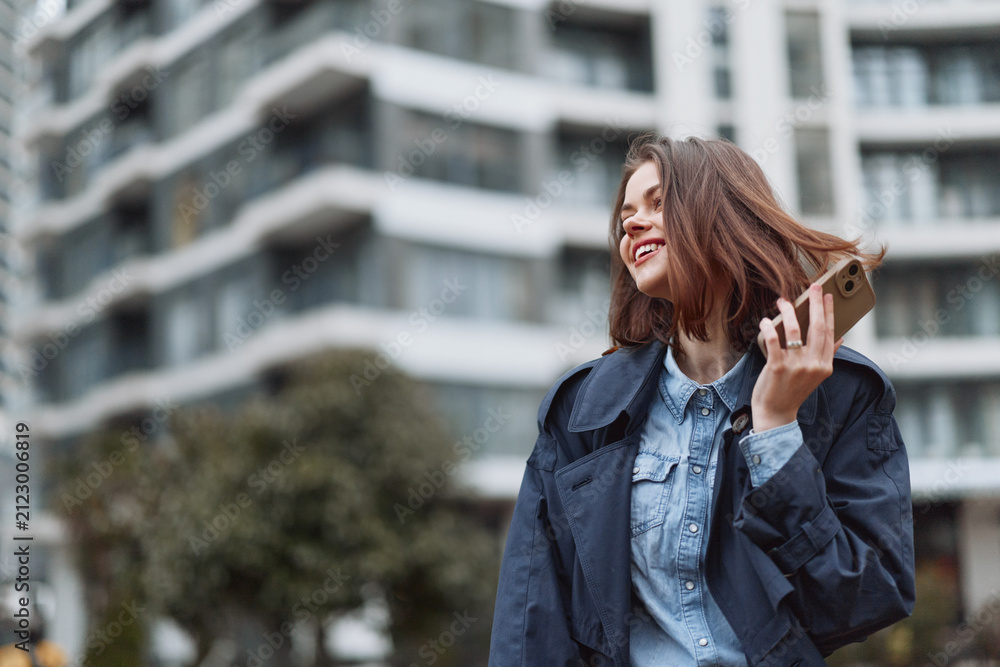 Fototapeta premium Young adult woman with brunette hair wearing a denim shirt and casual jacket talks on her smartphone in an urban street scene, smiling as modern architecture frames the shot.