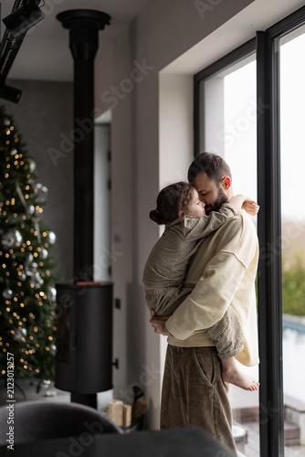 25.03.25 Bar, Ukraine: Man holds child near window as Christmas tree glows warmly inside cozy home