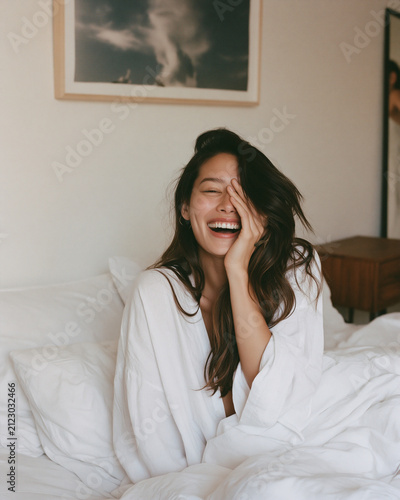 A beautiful woman seated on a bed in a well-lit room. She is laughing, with one hand covering her face, revealing her teeth