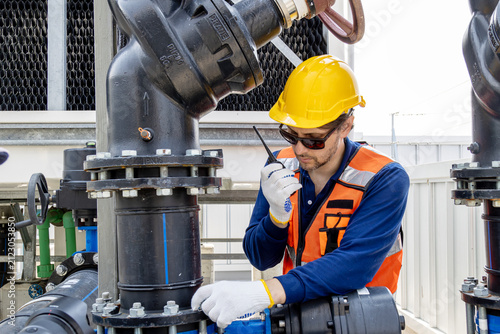 Industrial engineer with radio communicator inspecting factory cooling tower pipelineMaintenance technician in a hard hat and safety vest checking water valve pipesCaucasian supervisor monitoring.