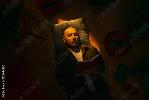 Top view of a relaxed bearded man lying on a wooden floor, reading an open red book under a warm beam of light in a dark, cozy room. Perfect evening for literature, storytelling and relaxation.