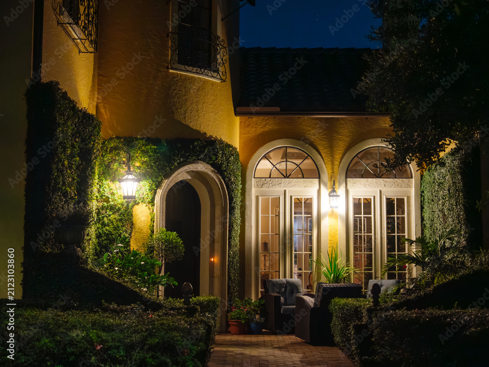 Fototapeta premium A beautifully illuminated courtyard entrance features arched doorways, landscaped hedges, and cozy outdoor seating. Warm lighting highlights the timeless architecture of the luxury residence after
