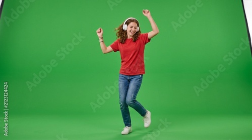 Cheerful teenage girl in red t-shirt and jeans dancing joyfully with white over-ear headphones against a bright green screen studio backdrop expressing happiness and youthful energy