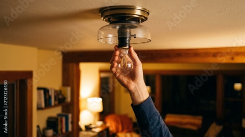 Hand screwing warm clear Edison bulb into vintage brass ceiling fixture in cozy wooden cabin interior with warm lamp glow, nighttime home maintenance and DIY repair concept