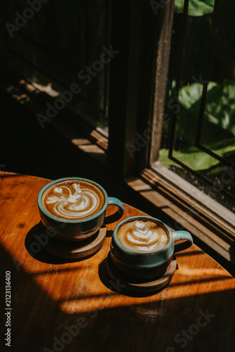 Moody aesthetic coffee photography showcasing two latte cups on wooden coasters, illuminated by strong window light and sharp shadows