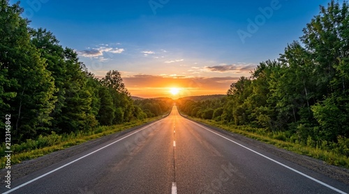 Empty straight asphalt road stretching toward glowing sunrise on horizon, flanked by lush green forest trees at dawn with vibrant orange sky and sunlit country highway perspective