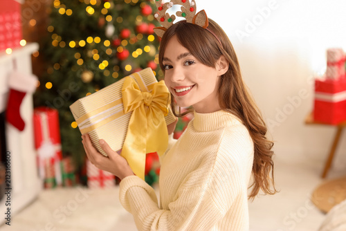 Beautiful young happy woman in reindeer horns with Christmas gift box near fi...