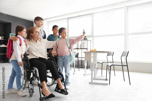 Mature teacher in wheelchair with little pupils taking selfie at school