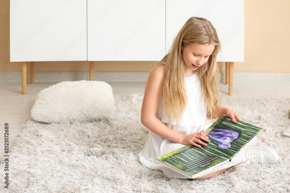 Naklejka premium Little girl reading book while sitting on floor at home