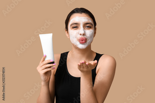 Beautiful young woman with facial mask blowing kiss on color background