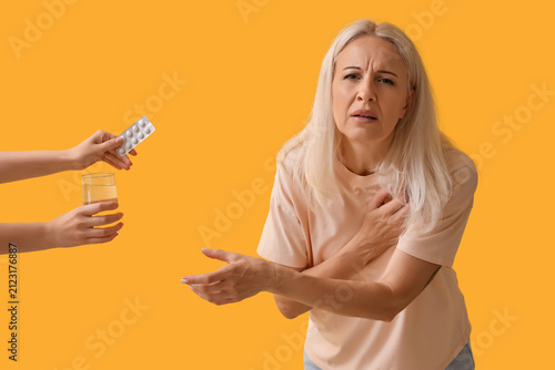 Mature woman with heart attack receiving pills and glass of water on yellow b...