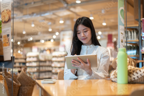 Fotografía Young asian woman writing in tablet while sitting with a bag and tumbler at tabl