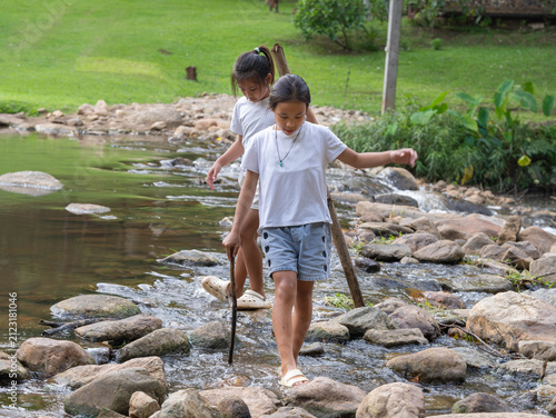 Two young Asian girls walking together across a shallow rocky stream in a lush green park during summer vacation