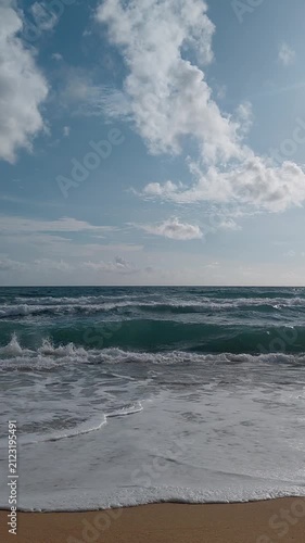 Soft turquoise sea ocean waves gently rolling onto a clean sandy beach shore under a bright blue sky with white clouds on a sunny day.
