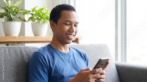Smiling happy young Indonesian man holding cell mobile phone using smartphone sitting at home on couch, scrolling social media, checking financial apps, buying online, texting messages. 