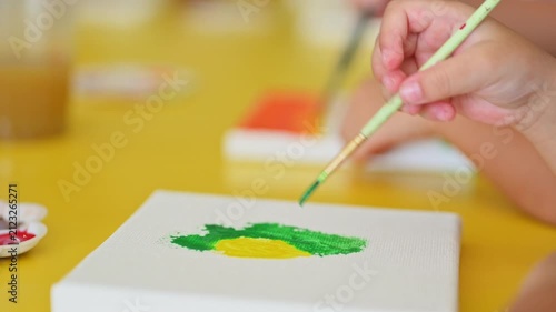 Child's Artistic Endeavor: A child's hand, holding a paintbrush, delicately adds strokes of vibrant green paint to a canvas, embarking on a creative painting activity.