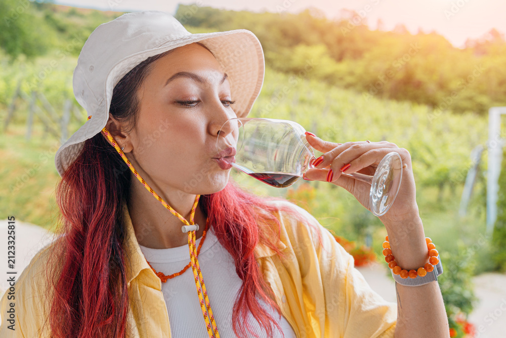 Obraz premium Young woman with pink hair sipping red wine at a sunny vineyard. Degustation tourism concept.