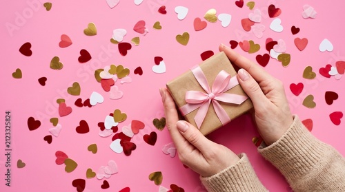 Hands holding a gift box with a pink ribbon on a pink background with heartshaped confetti