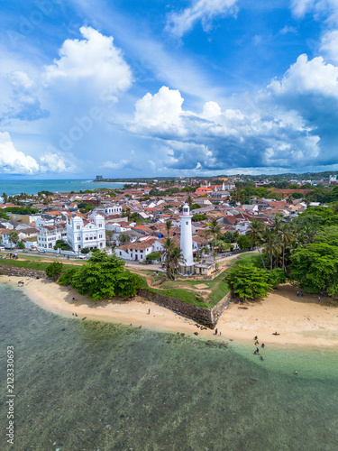 Vertical drone photo showing the tip of Galle Fort ramparts, Galle Lighthouse, pristine clear shallow coral waters, and the historic Dutch colonial townscape under a blue cloudy sky.