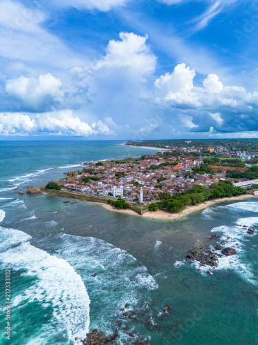 High-angle vertical aerial photograph showcasing the full layout of Galle Fort surrounded by turquoise coastal waters, crashing surf, and dramatic cloudy skies in Southern Sri Lanka.