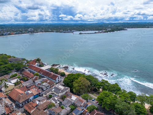 Drone photo looking across the coastal edge of Galle Fort, featuring the long white Old Dutch Hospital building, green trees, crashing waves, and the serene Galle Harbor Bay in Sri Lanka.