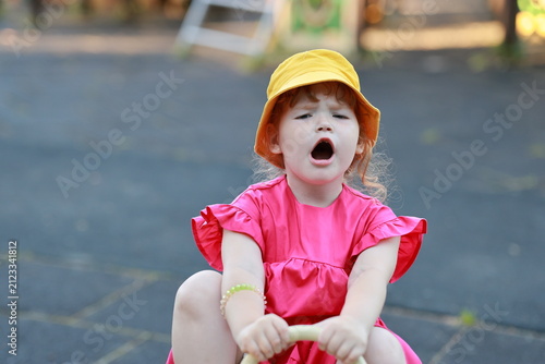 Child girl in a yellow hat playing on the playground. Cute kid in a pink dress having fun outdoors. Summer childhood leisure, active lifestyle, expression of joy and surprise.