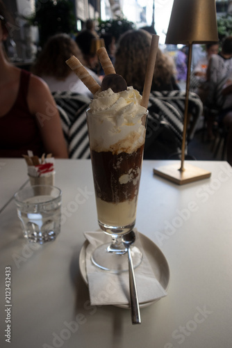 Elegante coppa di gelato affogato al caffè con strati di crema, cioccolato, panna montata e cialde croccanti, servita su un tavolo da bar all'aperto con lampada di design e atmosfera estiva.