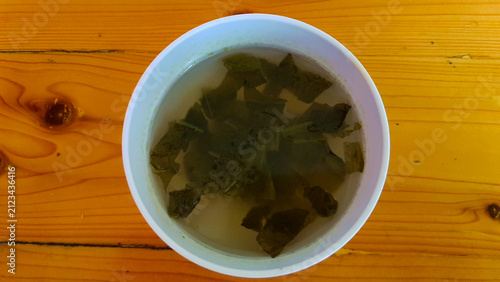 A top-down close-up photograph of fresh mustard greens soaking in water inside a white bowl, placed on a warm wooden table.